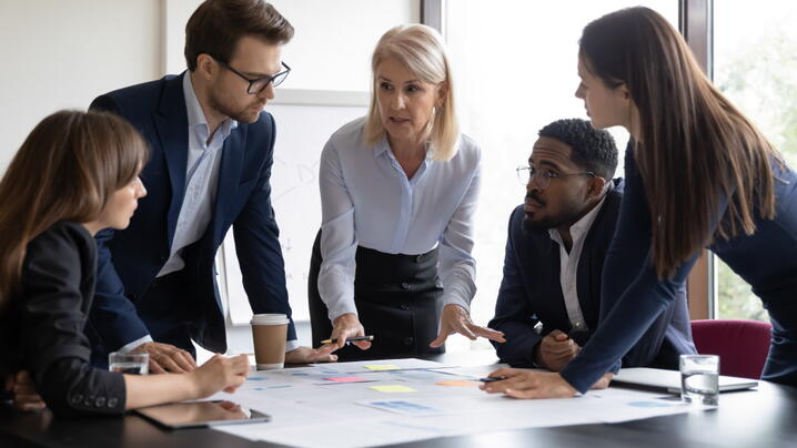 young businesspeople listening attentively to senior female leader discussing business plan at table